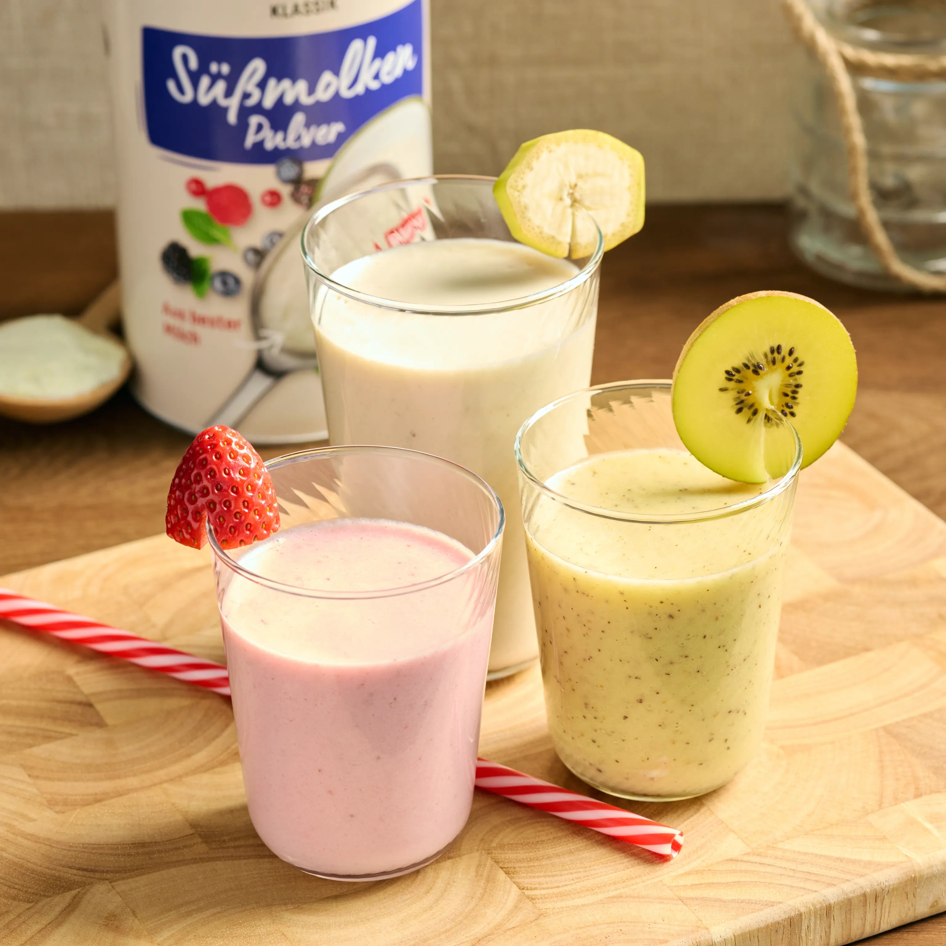 Drei Gläser mit Fruchtshakes auf Holzbrett, dekoriert mit Erdbeere, Kiwi und Banane, daneben Süßmolkenpulver.