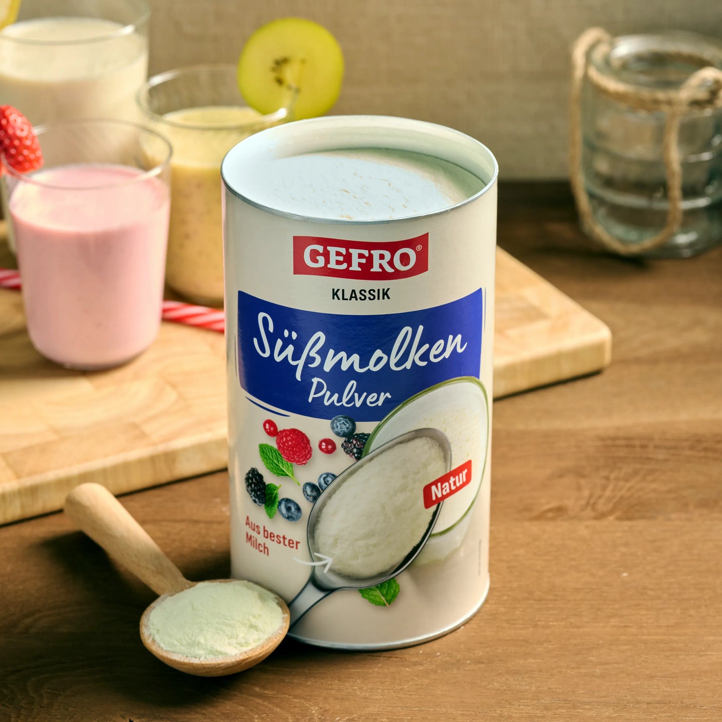 Dose GEFRO Süßmolkenpulver Natur auf Holztisch, davor ein Löffel Pulver, im Hintergrund Gläser mit Shakes.