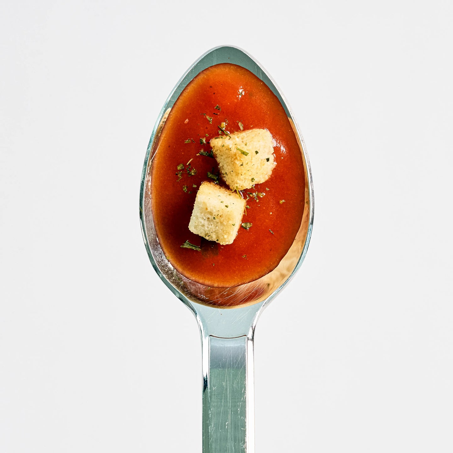 Löffel mit roter Tomatensuppe, garniert mit zwei Croutons und Kräutern, auf hellem Hintergrund von oben fotografiert