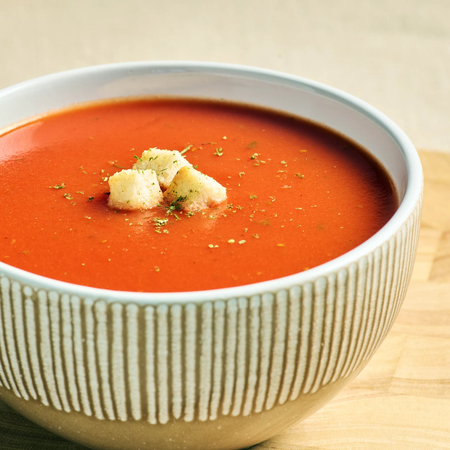 Schüssel mit cremiger Tomatensuppe, garniert mit Croutons und Kräutern, servierfertig auf hellem Holztisch.