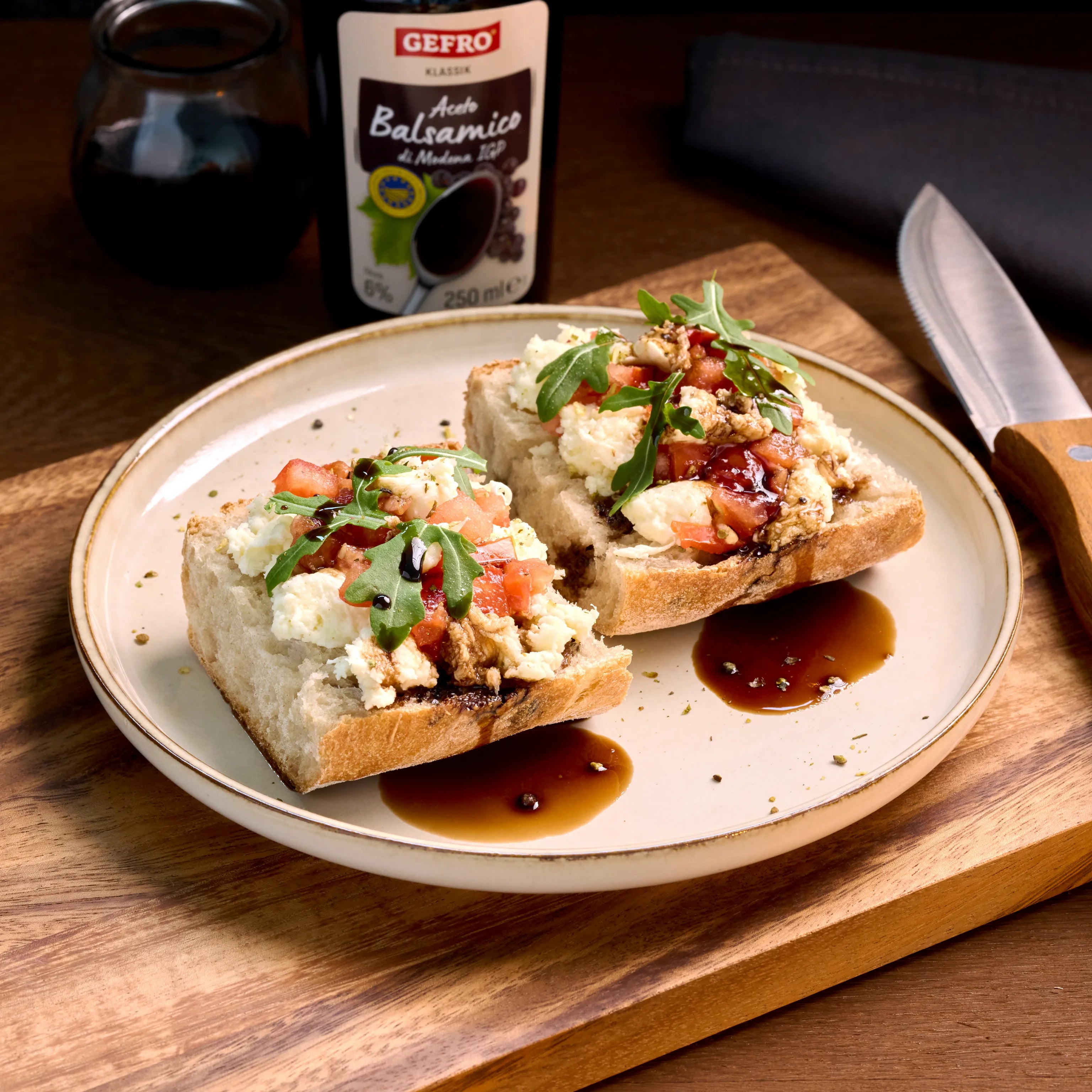 Zwei belegte Brotscheiben mit Käse, Tomaten, Rucola und Balsamico auf einem Teller, daneben eine Balsamico-Flasche.