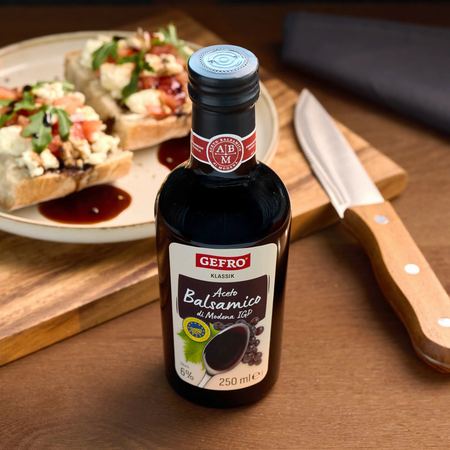 Flasche GEFRO Aceto Balsamico di Modena auf Holztisch, daneben belegte Brote und Messer als Serviervorschlag
