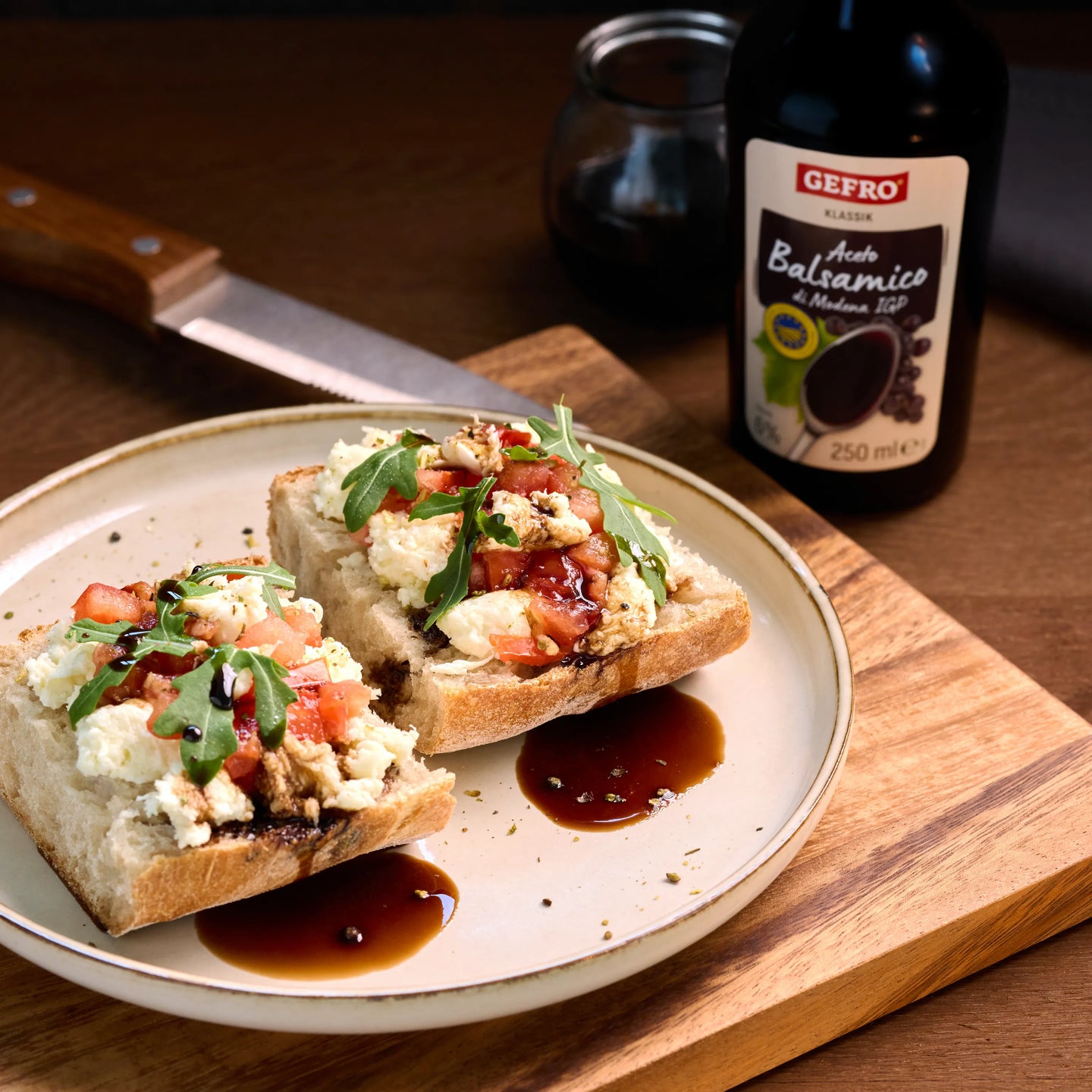 Zwei belegte Brote mit Käse, Tomaten und Rucola auf einem Teller, daneben eine Flasche Aceto Balsamico.