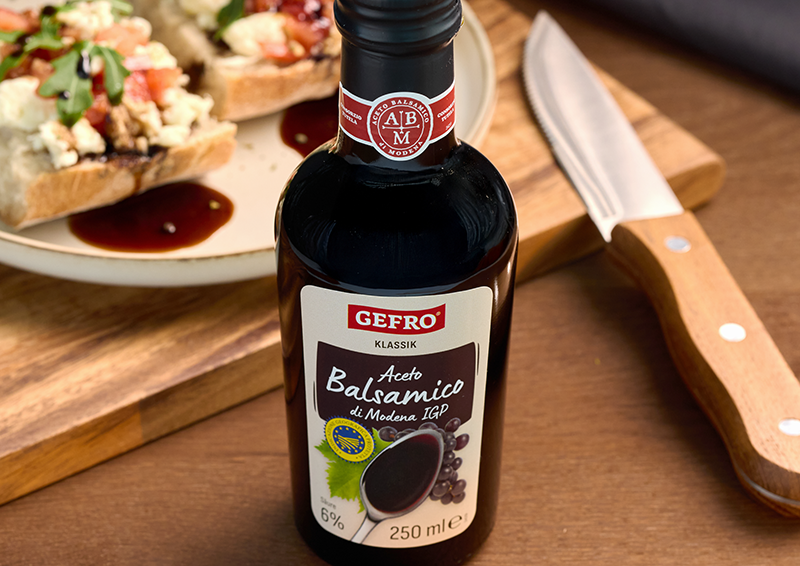 Flasche Gefro Aceto Balsamico di Modena auf Holztisch, daneben Bruschetta auf Teller und ein Messer.