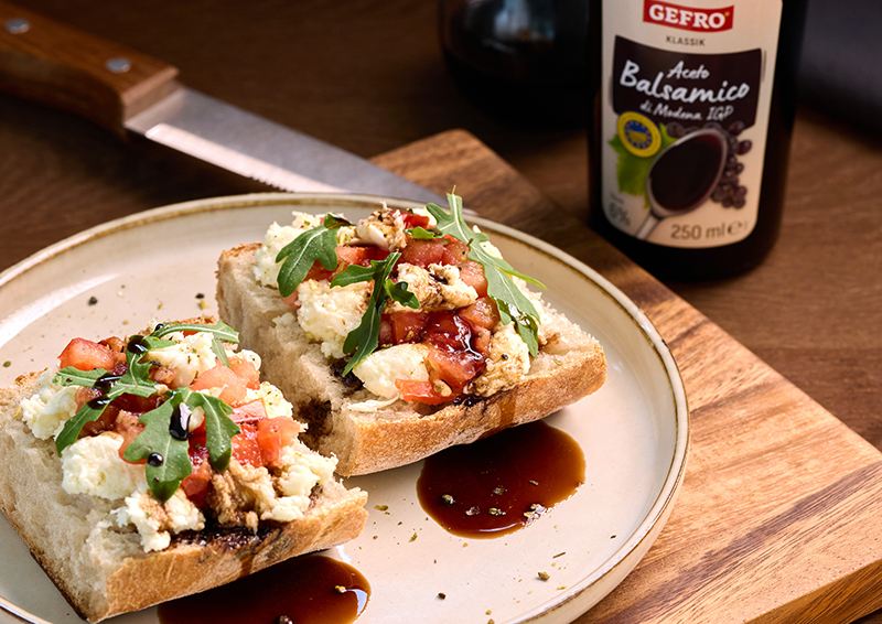 Belegte Brote mit Käse, Tomaten und Rucola auf einem Teller, daneben Balsamico-Essig auf Holzbrett