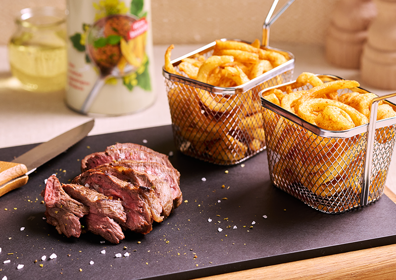 Geschnittenes Steak auf schwarzer Platte mit grobem Salz, dazu zwei Körbe mit Pommes im warmen Küchenlicht