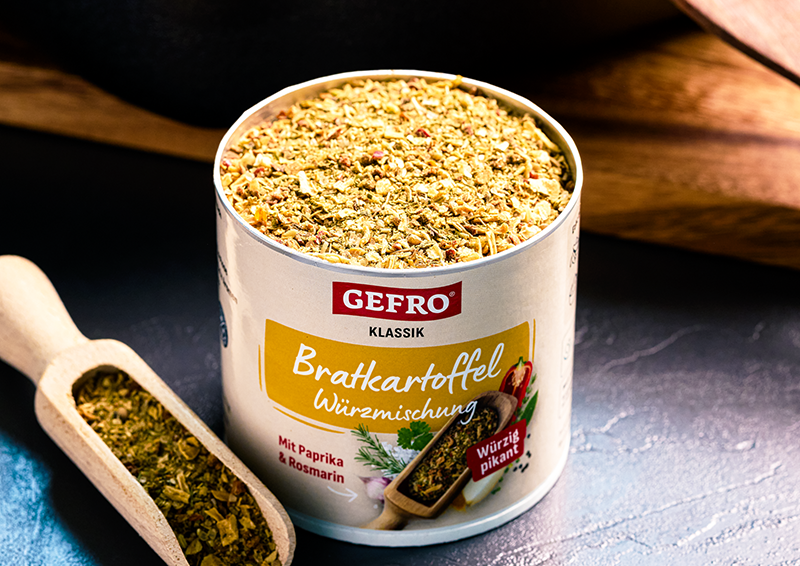 Dose mit GEFRO Bratkartoffel Würzmischung, offen gezeigt mit Kräutern und Gewürzen, daneben eine kleine Holzschaufel