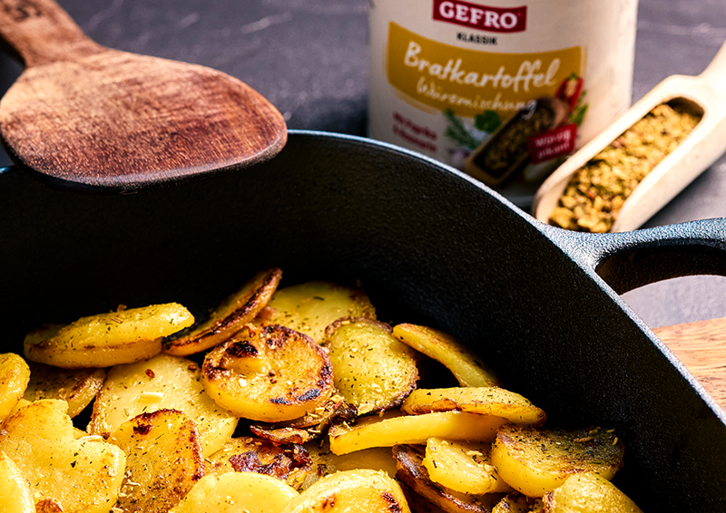 Gebratene Kartoffelscheiben in einer Pfanne, daneben Holzlöffel und Bratkartoffel-Gewürz im Hintergrund