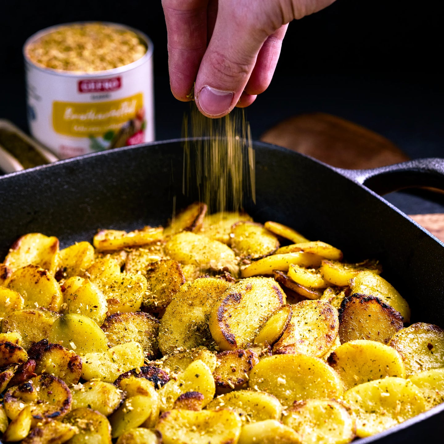 Hand würzt goldbraune Bratkartoffeln in einer Pfanne, im Hintergrund steht eine Dose Kartoffelgewürz.