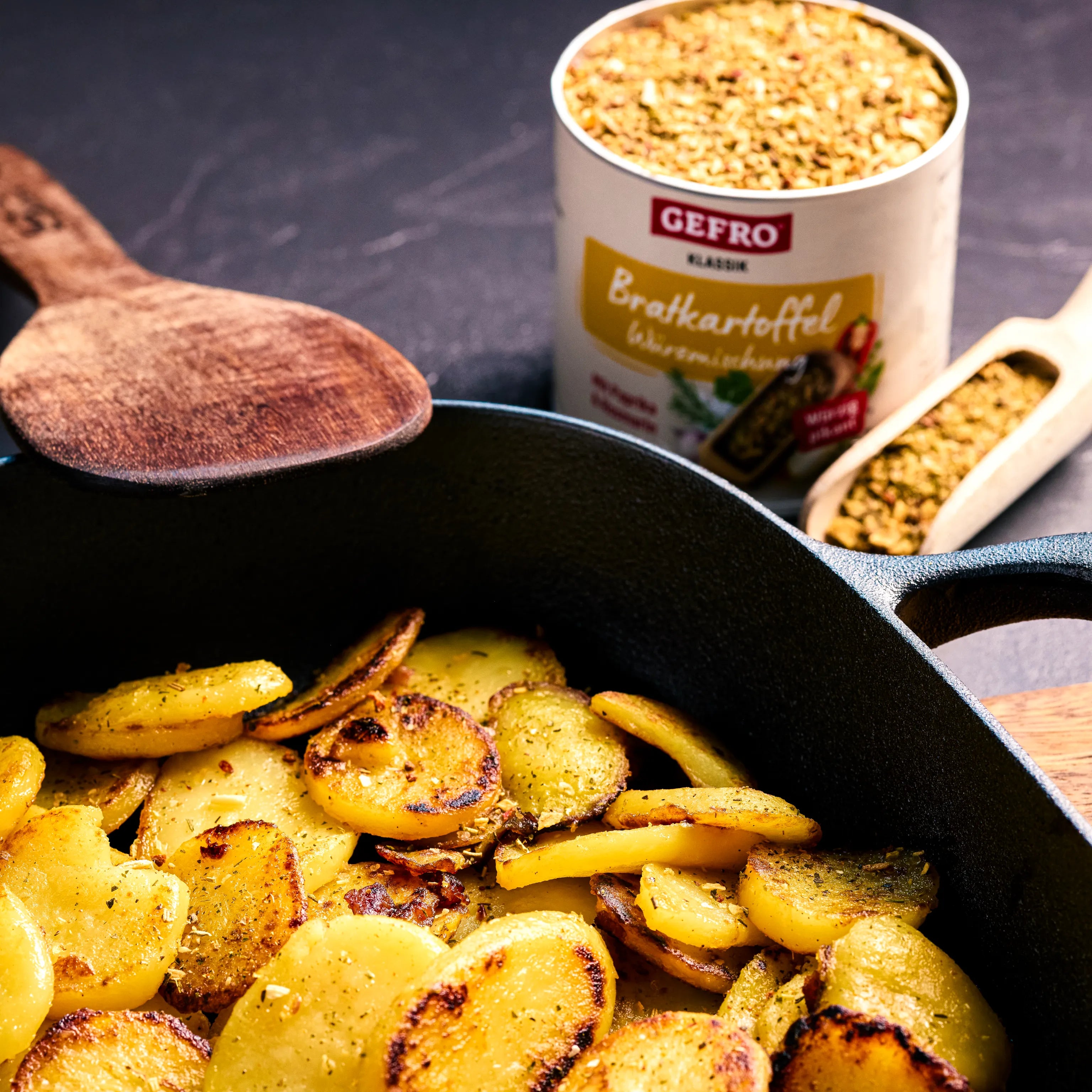 Gebratene Kartoffelscheiben in einer Pfanne, daneben Gewürzmischung und Holzlöffel zur Zubereitung von Bratkartoffeln