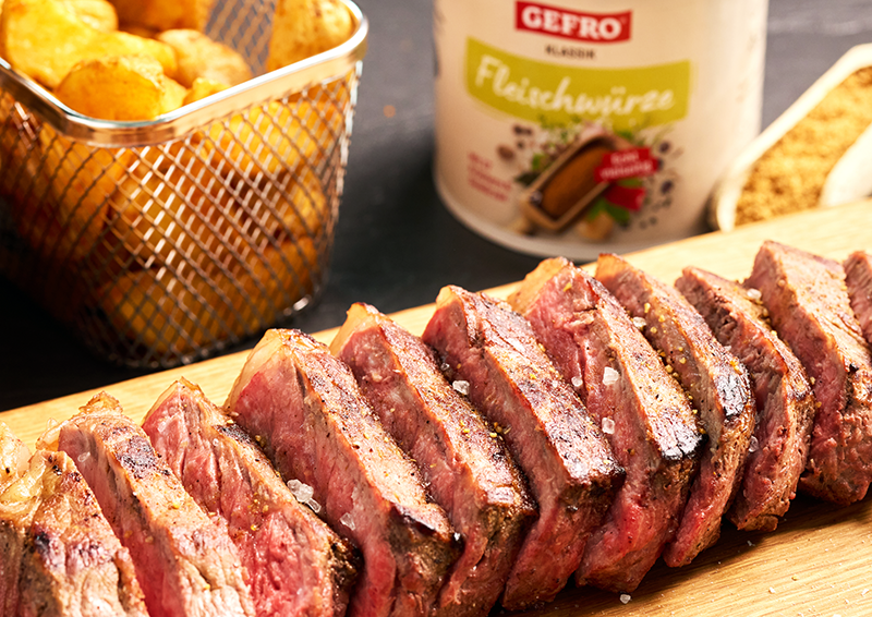 Aufgeschnittenes Steak auf Holzbrett mit grobem Salz, dazu Kartoffelbeilagen und Fleischwürze im Hintergrund