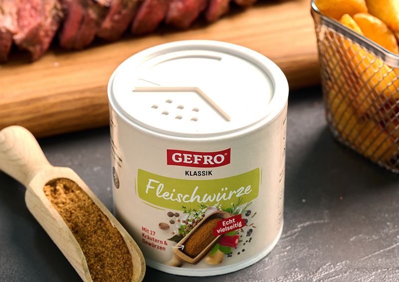 Dose GEFRO Klassik Fleischwürze auf Tisch, daneben Gewürzschaufel, Steak im Hintergrund und Korb mit Chips