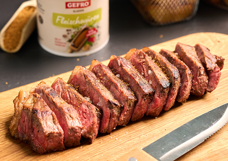 In Scheiben geschnittenes, rosa gebratenes Steak auf Holzbrett mit Messer, im Hintergrund Gewürzdose.