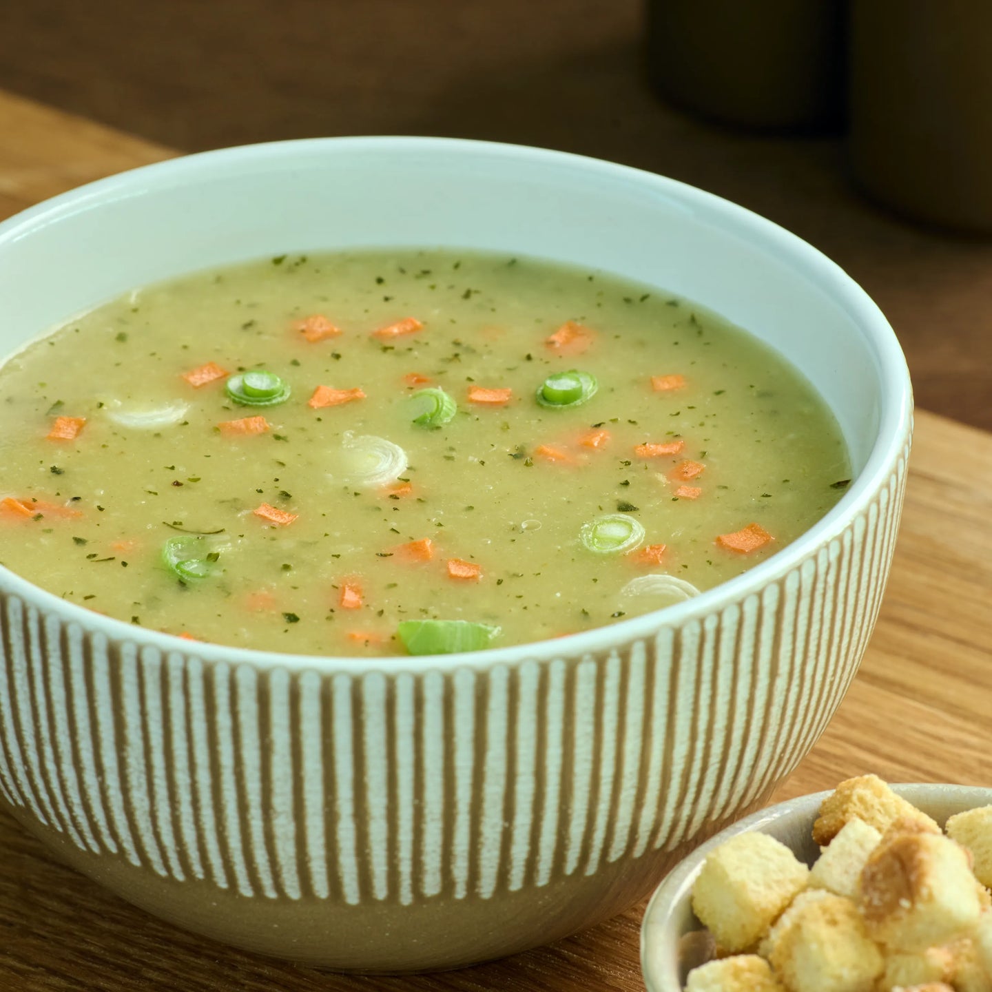 Schüssel mit cremiger Gemüsesuppe, garniert mit Frühlingszwiebeln und Karottenstückchen, auf Holztisch.