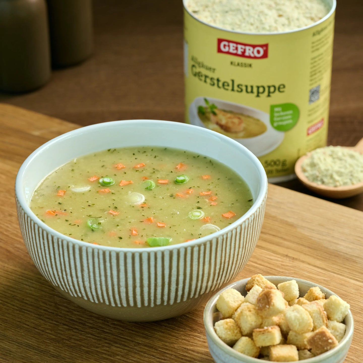 Schüssel mit heller Gerstelsuppe und Gemüsewürfeln auf Holzbrett, daneben Croutons und die GEFRO Packung im Hintergrund