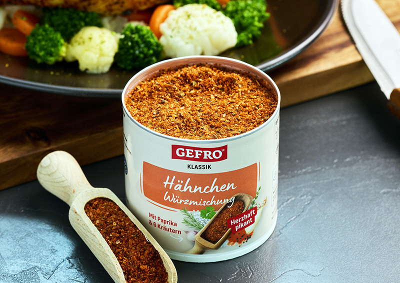 Dose Gefro Hähnchen-Würzmischung mit offenem Gewürz, Holzschaufel und Gemüsegericht im Hintergrund