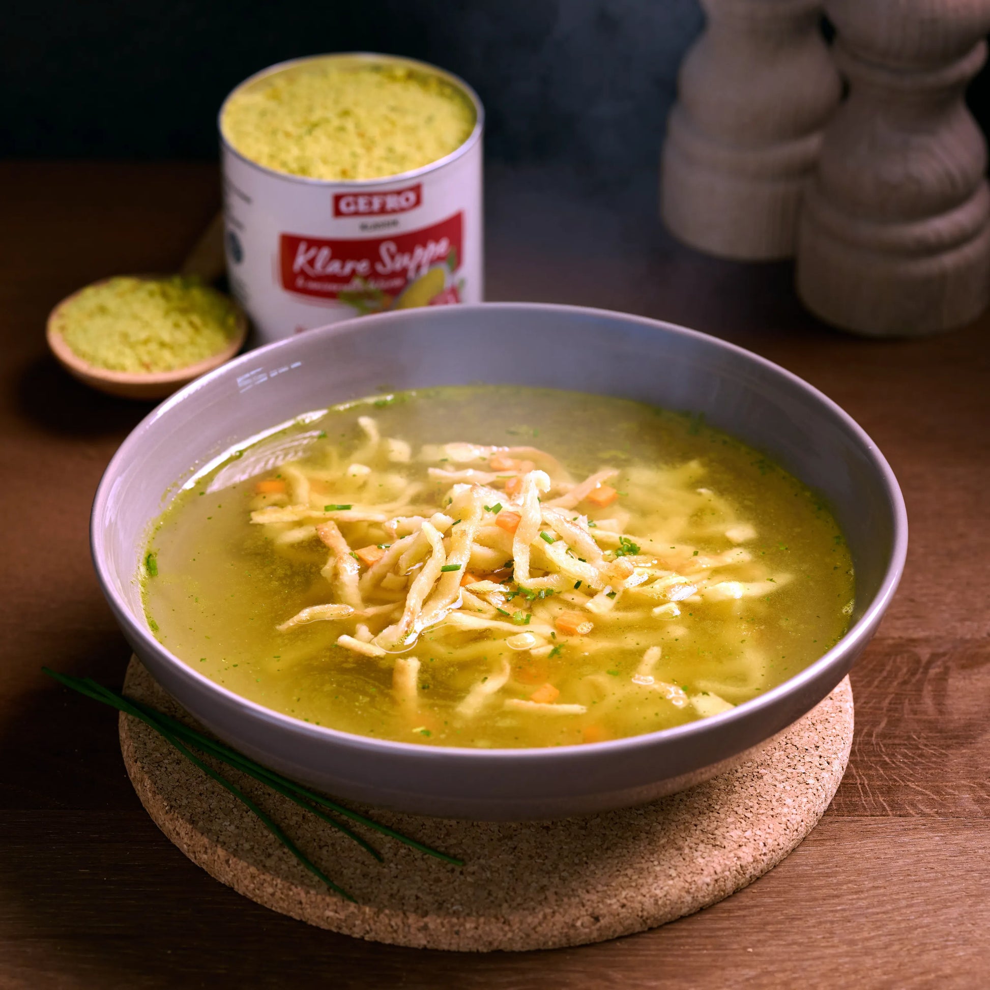 Schüssel mit klarer Nudelsuppe auf Holzunterlage, dahinter Dose Brühepulver und Löffel mit Granulat