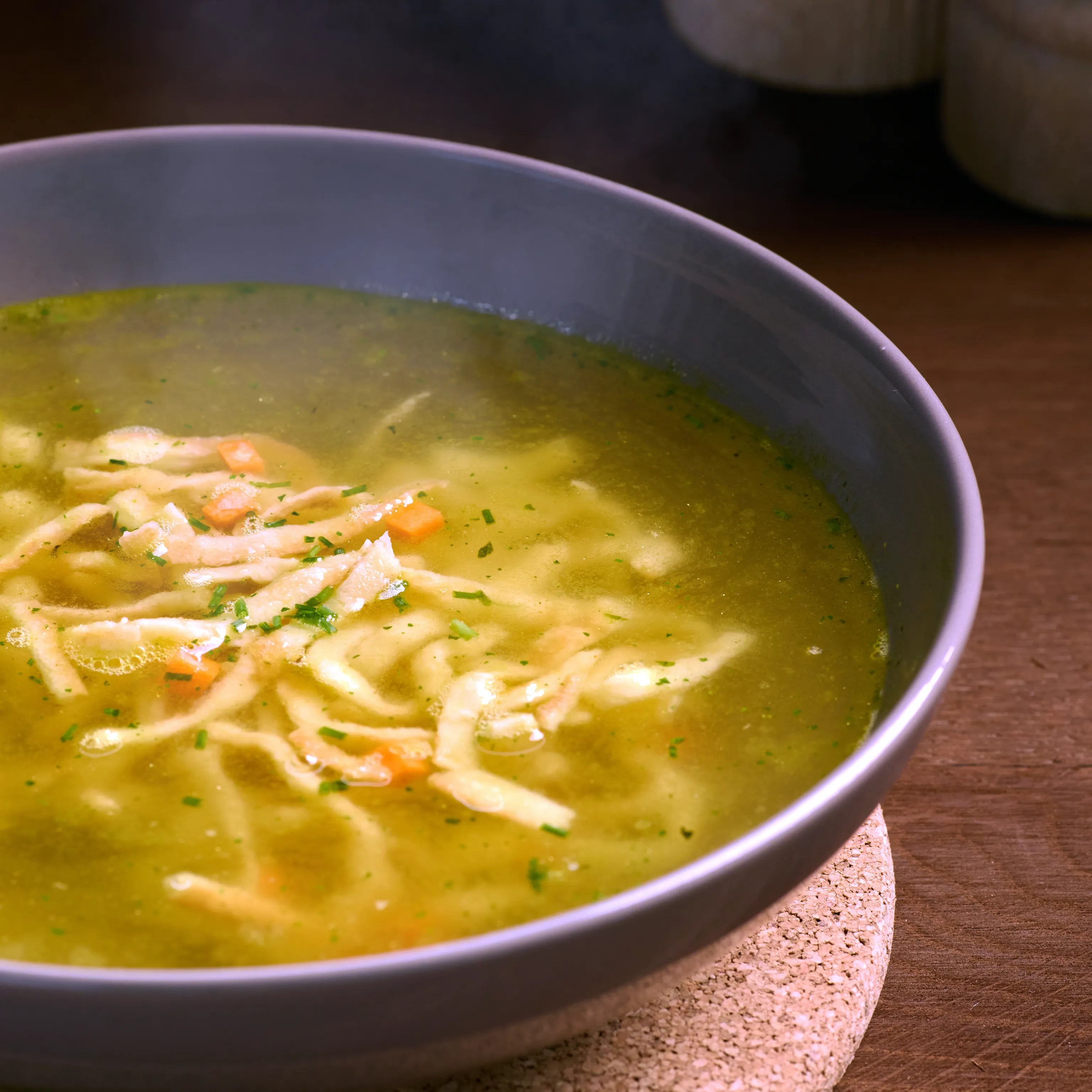 Graue Schale mit klarer Nudelsuppe, feinen Gemüsestücken und Kräutern auf Holzuntergrund, warm serviert