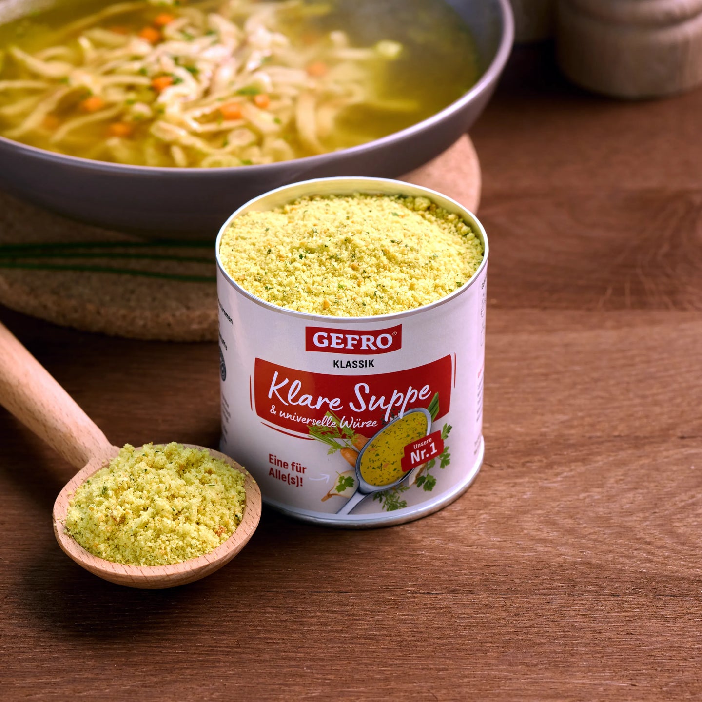 Dose GEFRO Klare Suppe mit Pulver im Holzlöffel, davor eine Schüssel Nudelsuppe auf Holztisch.