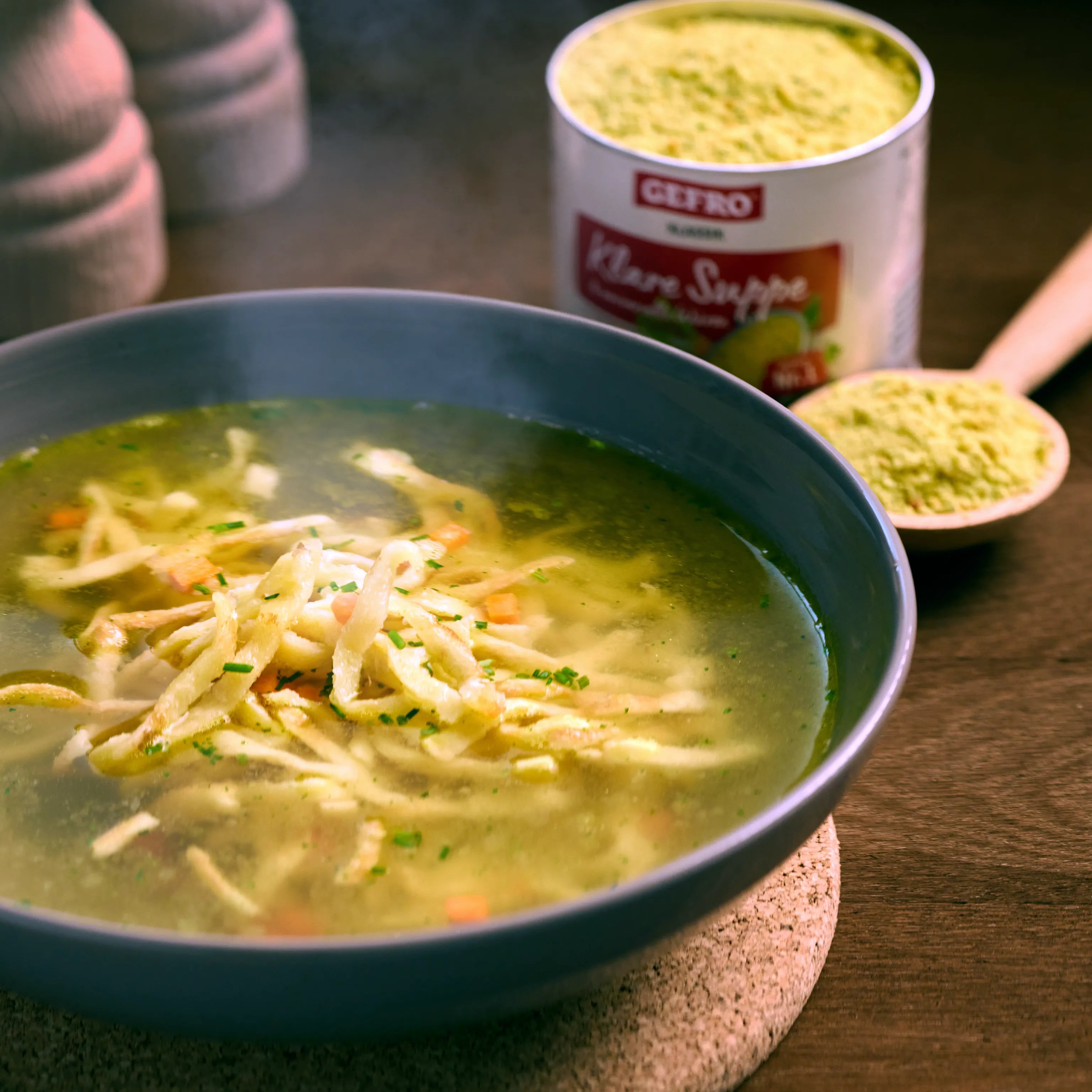 Schüssel mit klarer Suppe und feinen Nudeln auf Holztisch, daneben Becher und Löffel mit Hühnerbrühe-Pulver