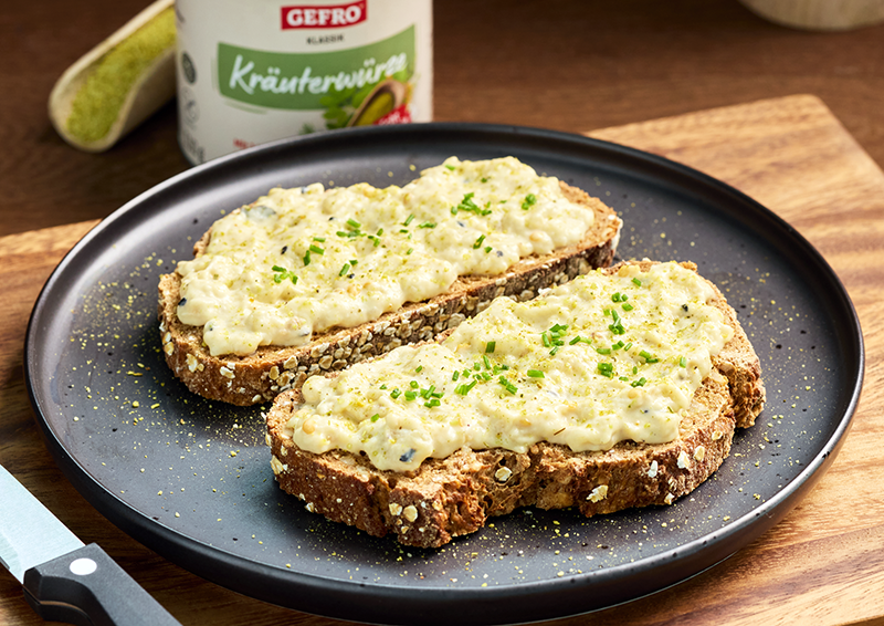 Zwei Scheiben Körnerbrot mit cremigem Eiersalat auf schwarzem Teller, mit Schnittlauch und Gewürz bestreut