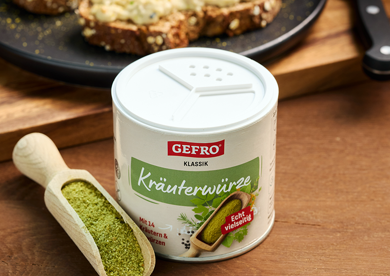 Dose GEFRO Kräuterwürze auf Holztisch mit gefüllter Holzschaufel, im Hintergrund belegte Brote auf einem Teller