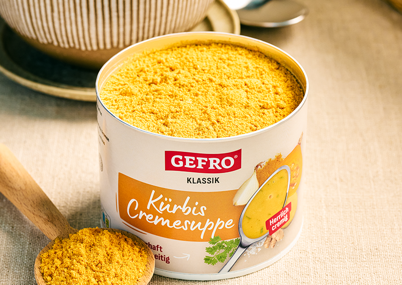 Dose Gefro Klassik Kürbis Cremesuppe mit geöffnetem Pulverinhalt auf Tisch, daneben ein Holzlöffel mit Suppepulver