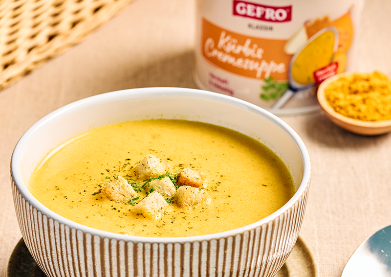 Schüssel mit cremiger Kürbis-Cremesuppe und Croûtons, daneben Verpackung und Gewürzschale auf hellem Tisch