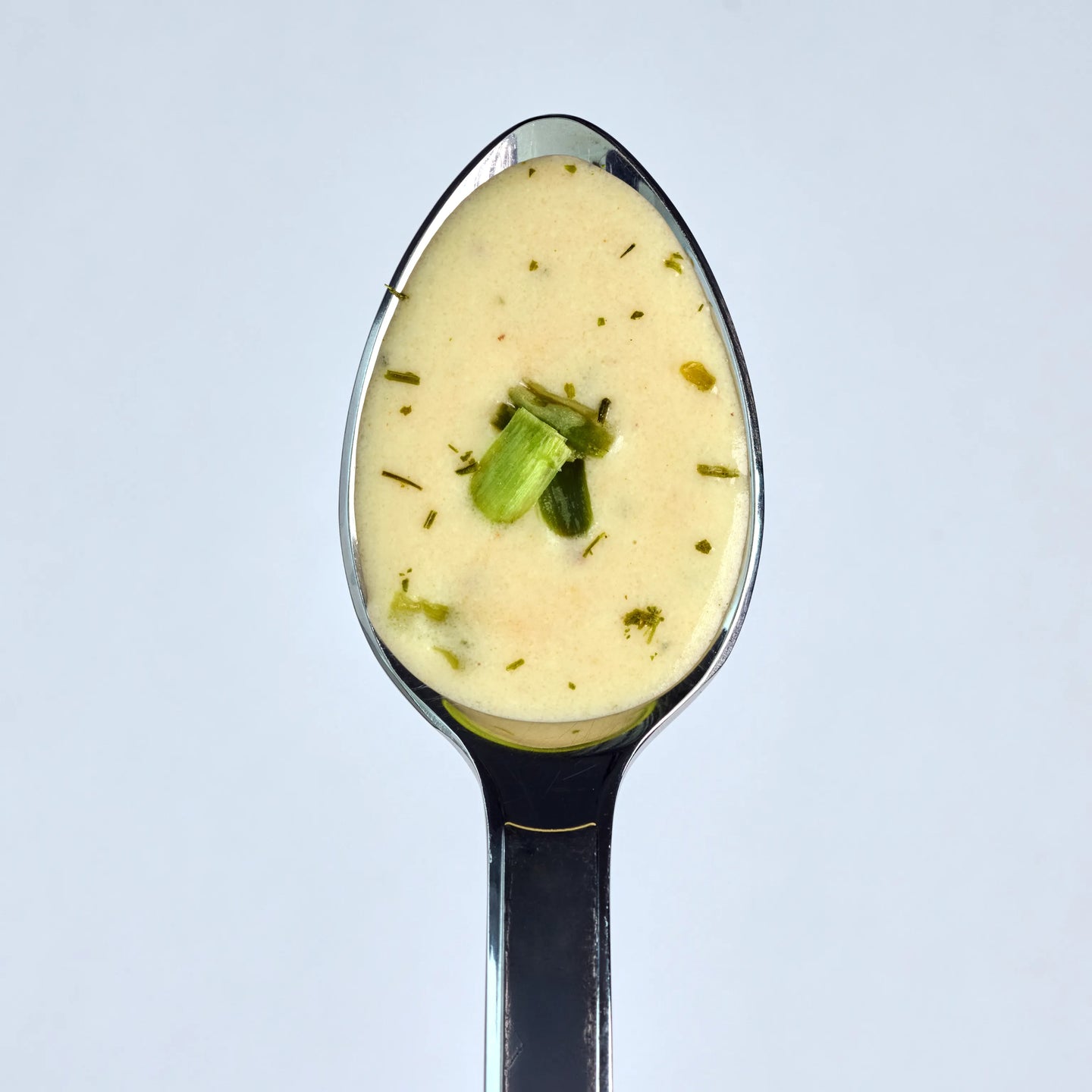 Löffel mit cremiger, heller Suppe oder Sauce mit Kräutern und kleinen Frühlingszwiebelstücken vor hellem Hintergrund