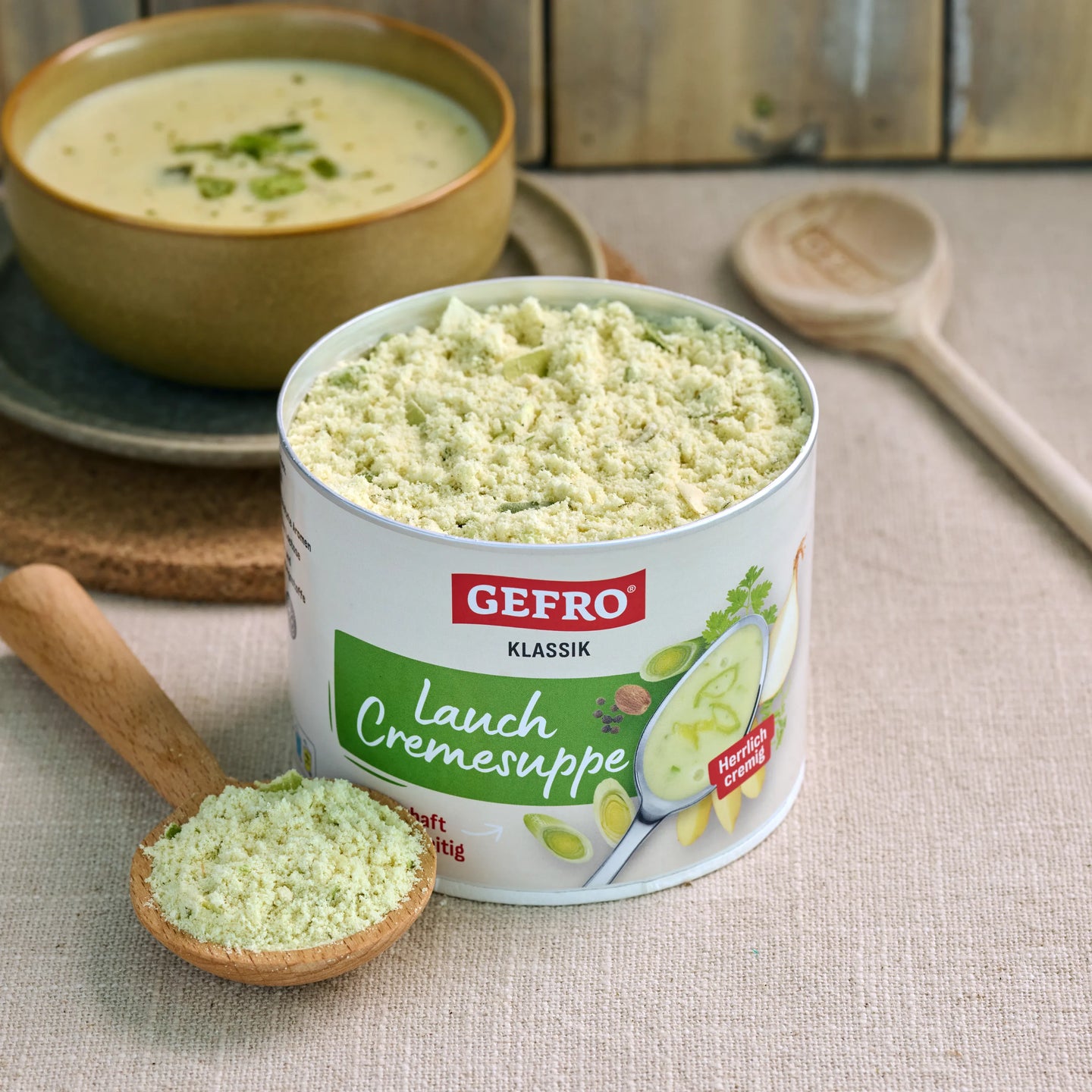 Dose GEFRO Klassik Lauch Cremesuppe mit Suppenpulver im Holzlöffel, dahinter eine angerichtete Schüssel Suppe