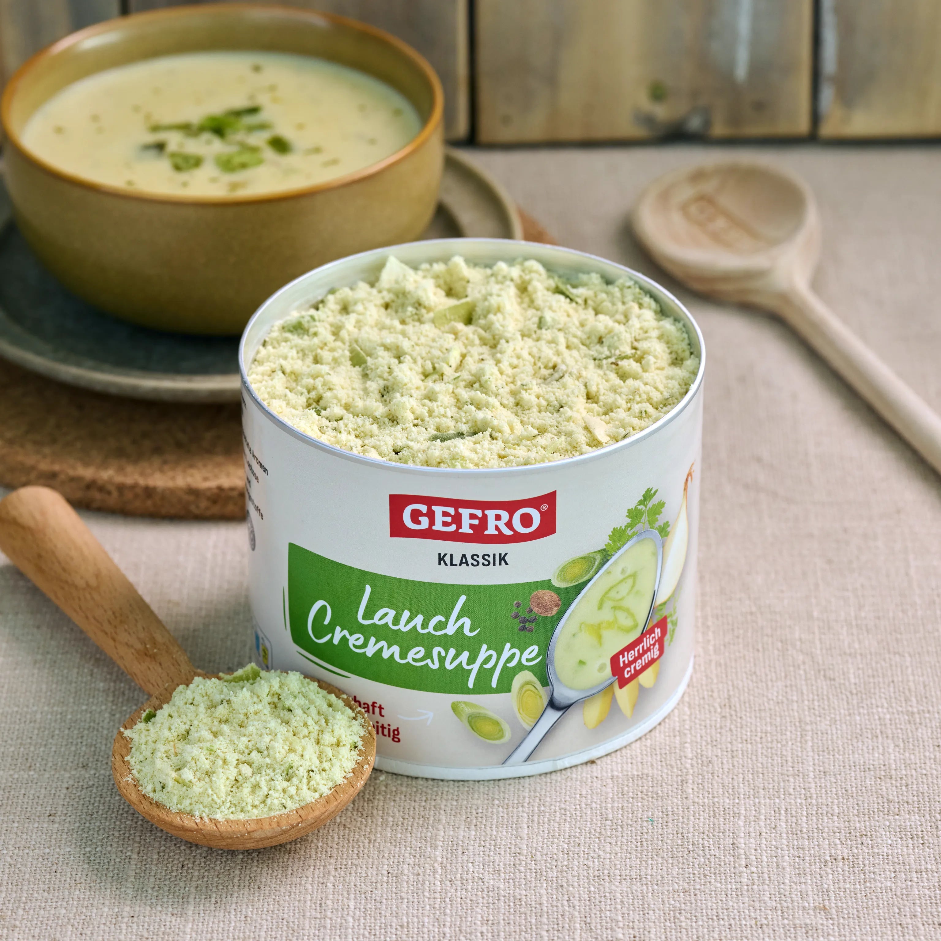 Dose GEFRO Klassik Lauch Cremesuppe mit Suppenpulver im Holzlöffel, dahinter eine angerichtete Schüssel Suppe
