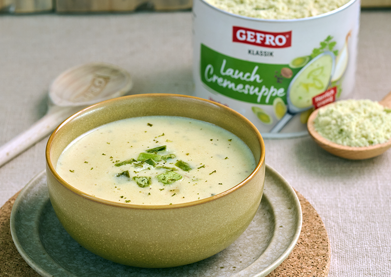 Schale mit cremiger Lauchsuppe auf Teller, daneben GEFRO Klassik Lauch Cremesuppe und ein Löffel Suppenpulver