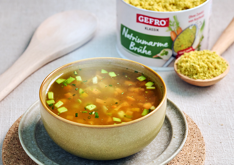 Schüssel mit klarer Gemüsesuppe auf Teller, daneben GEFRO Naturwürze Brühe und Holzlöffel auf hellem Tisch