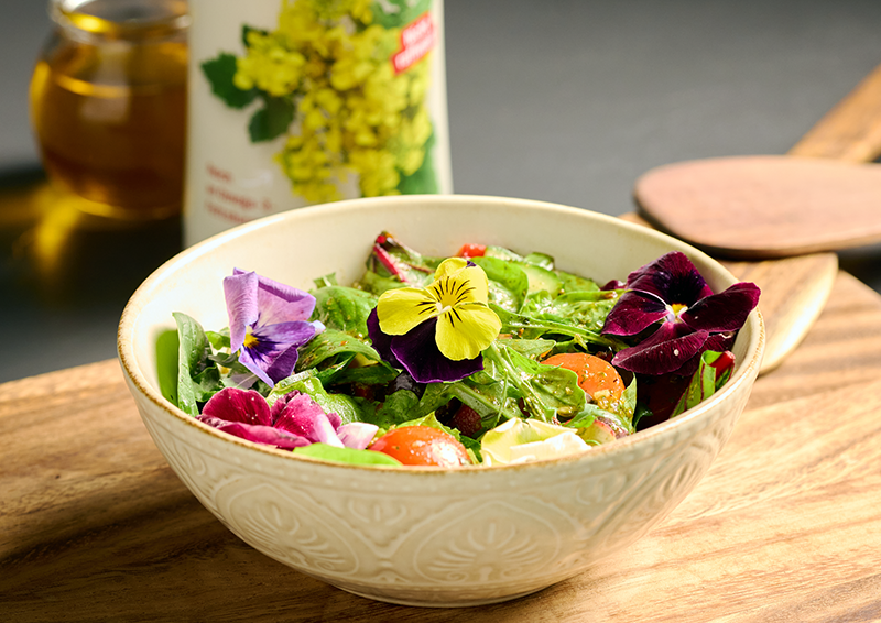 Schüssel mit frischem Salat, essbaren Blüten, Tomaten und Gurke auf Holztisch, daneben Ölflasche im Hintergrund