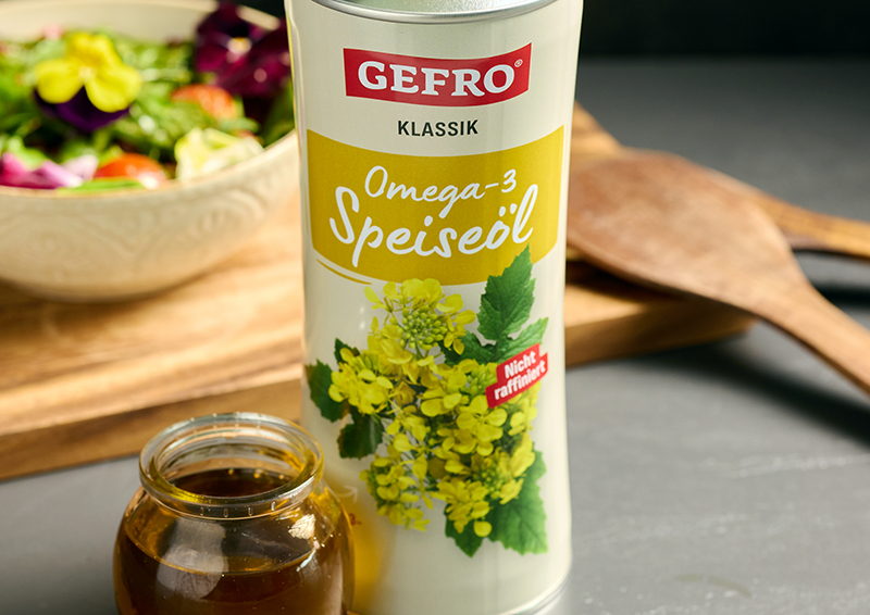 GEFRO Klassik Omega-3 Speiseöl in Dose neben kleinem Glas, dahinter Salatschüssel und Holzlöffel auf Tisch