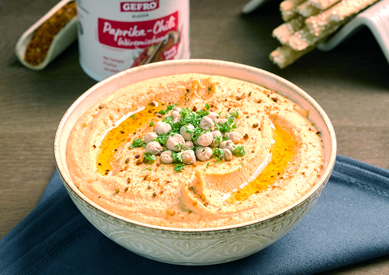 Schüssel mit cremigem Paprika-Chili-Hummus, garniert mit Kichererbsen, Petersilie und Öl auf dunklem Tuch.