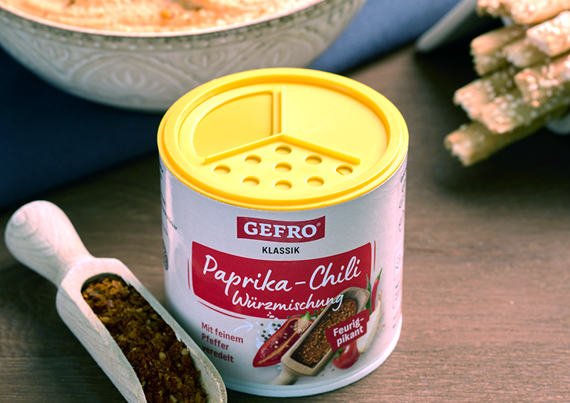 Dose mit Paprika-Chili-Würzmischung von Gefro auf dem Tisch, daneben Holzschaufel mit Gewürz und Schale im Hintergrund