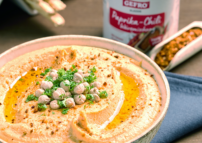 Schale mit cremigem Hummus, Kichererbsen, Petersilie und Öl, dazu Paprika-Chili-Gewürz im Hintergrund