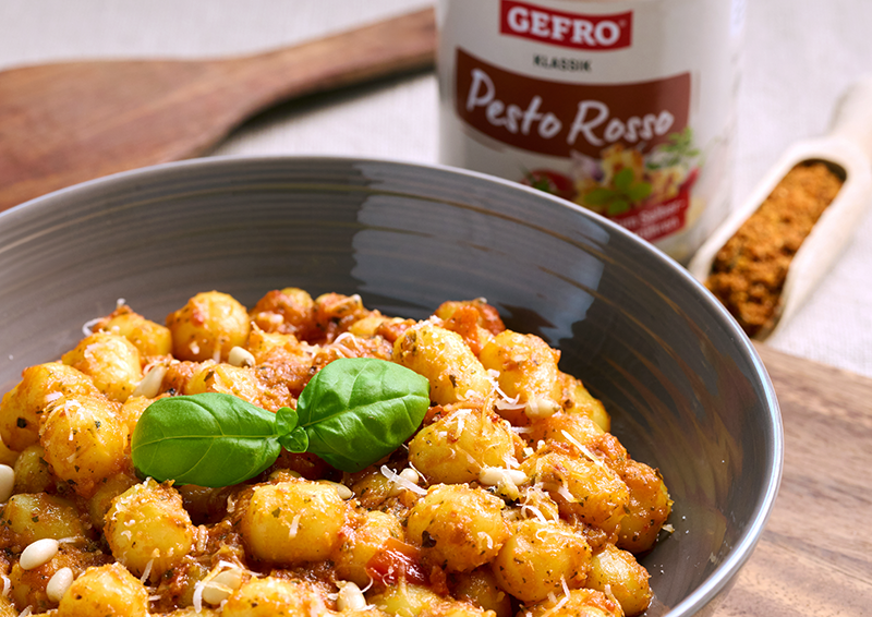 Schale mit Gnocchi in rotem Pesto, garniert mit Basilikum und Käse; im Hintergrund ein Glas Pesto Rosso.