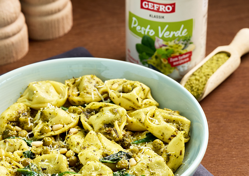 Schüssel mit Tortellini in grünem Pesto mit Erbsen und Käse, daneben ein Glas Pesto Verde auf dem Tisch