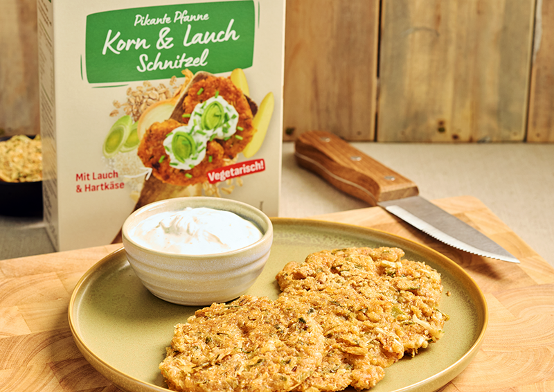 Zwei vegane Schnitzel auf Teller mit Dip, daneben Verpackung „Korn & Lauch Schnitzel“ auf Holztisch in Küche