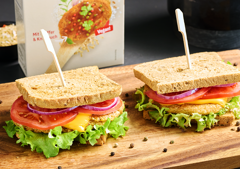 Zwei belegte Sandwiches mit Salat, Tomate, Zwiebeln, Käse und veganem Schnitzel auf einem Holzbrett.