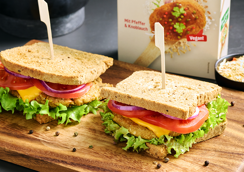 Zwei belegte vegane Sandwiches mit Salat, Tomate, Zwiebeln und Käseersatz auf einem Holzbrett angerichtet