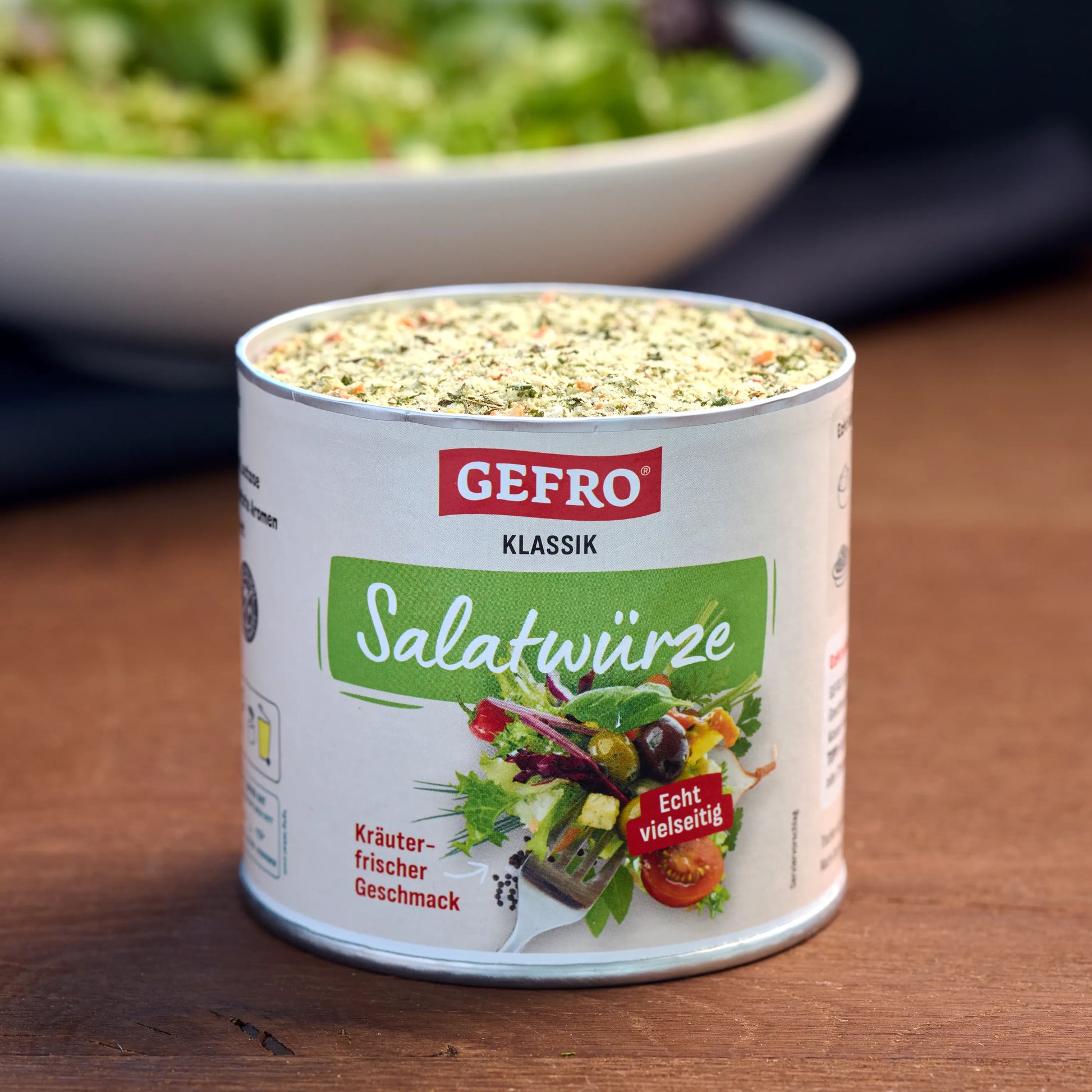 Dose GEFRO Klassik Salatwürze auf Holztisch, geöffnet mit sichtbarer Würzmischung, vor unscharfer Salatschüssel