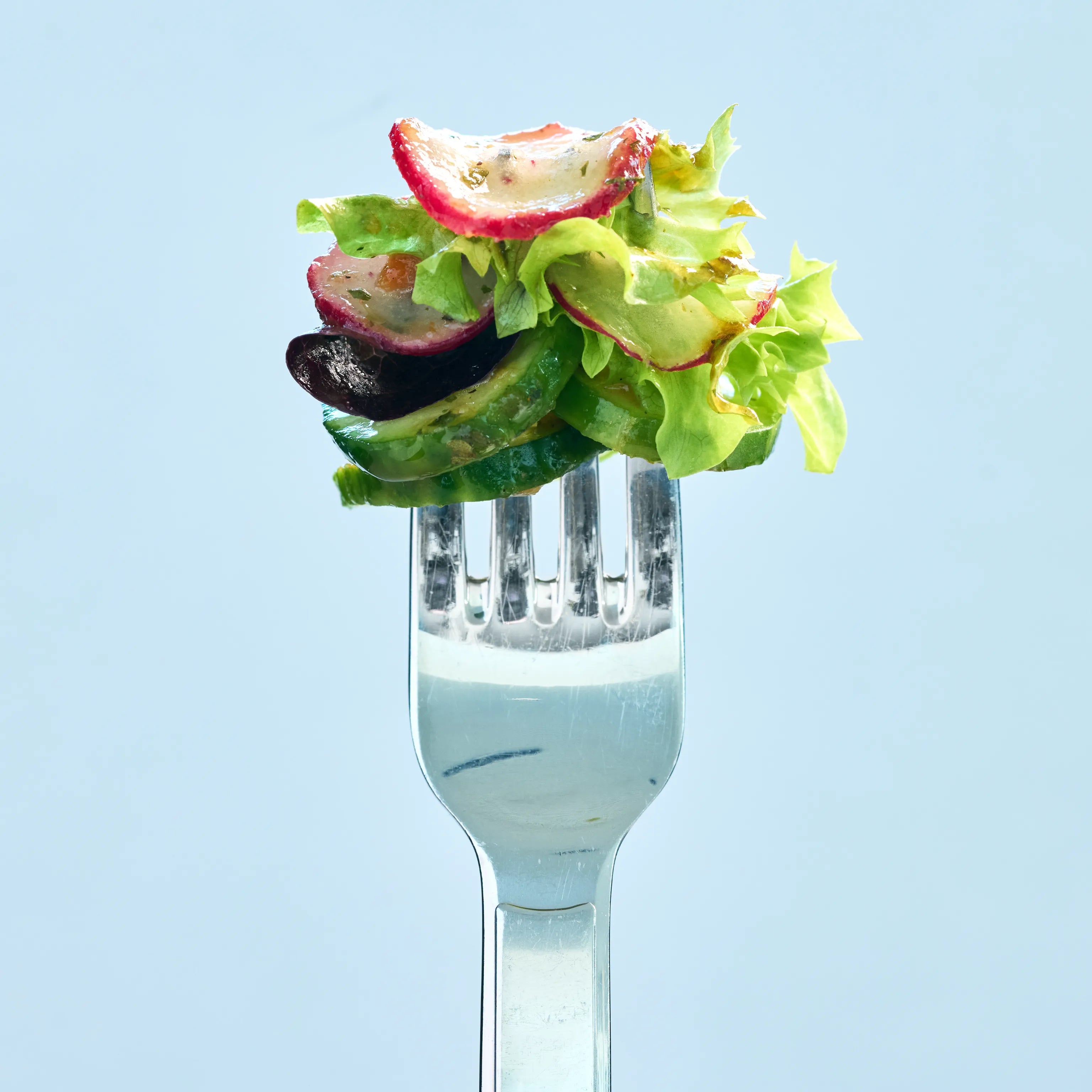 Gabel mit frischem Salat aus Gurke, Radieschen, Blattsalat und Olive vor hellem blauem Hintergrund