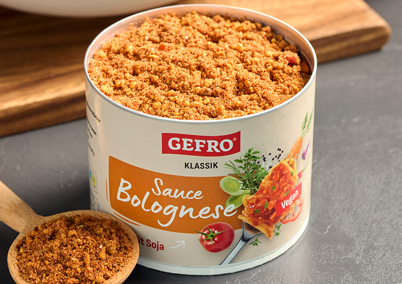 Dose GEFRO Klassik Sauce Bolognese mit Soja, geöffnet und randvoll mit Granulat, daneben ein Holzlöffel mit Inhalt