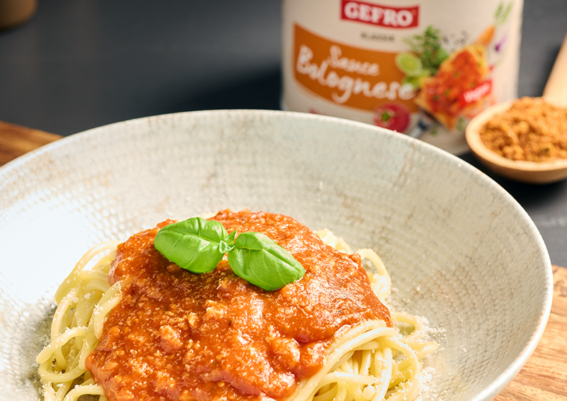 Teller mit Spaghetti und roter Bolognese, garniert mit Basilikum; im Hintergrund ein Gewürzglas und eine Schale```