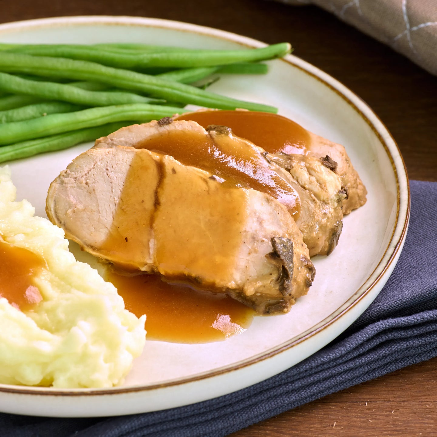 Aufgeschnittenes Schweinebratenstück mit brauner Soße, Kartoffelpüree und grünen Bohnen auf hellem Teller.