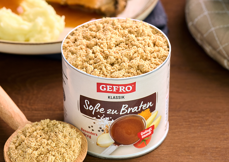 Dose GEFRO Klassik Soße zu Braten mit geöffnetem Pulverinhalt, davor ein Holzlöffel mit Soßenpulver auf dem Tisch