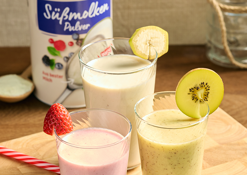 Drei Smoothies im Glas mit Erdbeere, Kiwi und Banane auf Holzbrett, daneben eine Packung Süßmolkenpulver